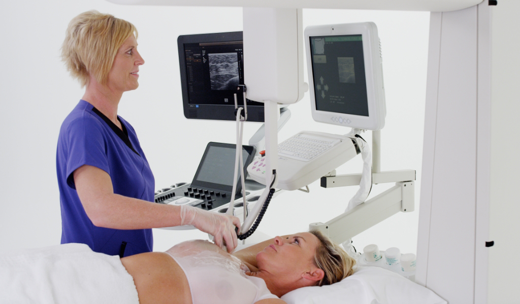 Imaging technician performing breast scan on female patient, with ultrasound results on screen
